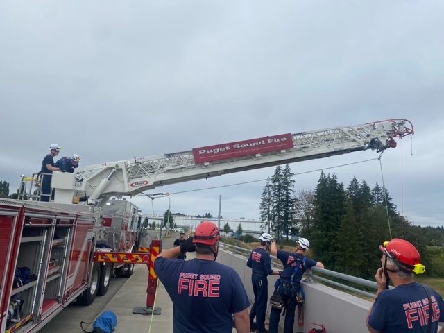 A group of firefighters stand on a bridge looking up at a crane attached to a firetruck